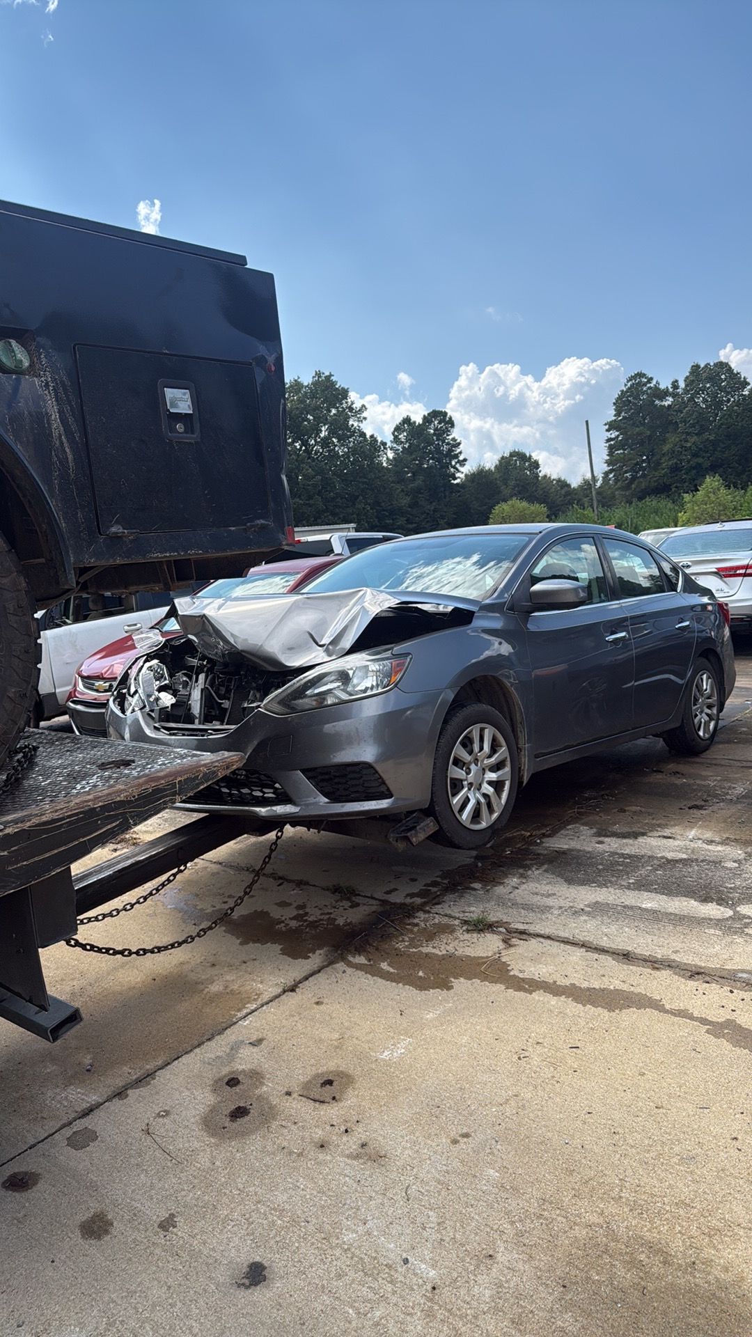 Gray sedan with heavy front-end damage on a tow truck in a parking lot under a blue sky