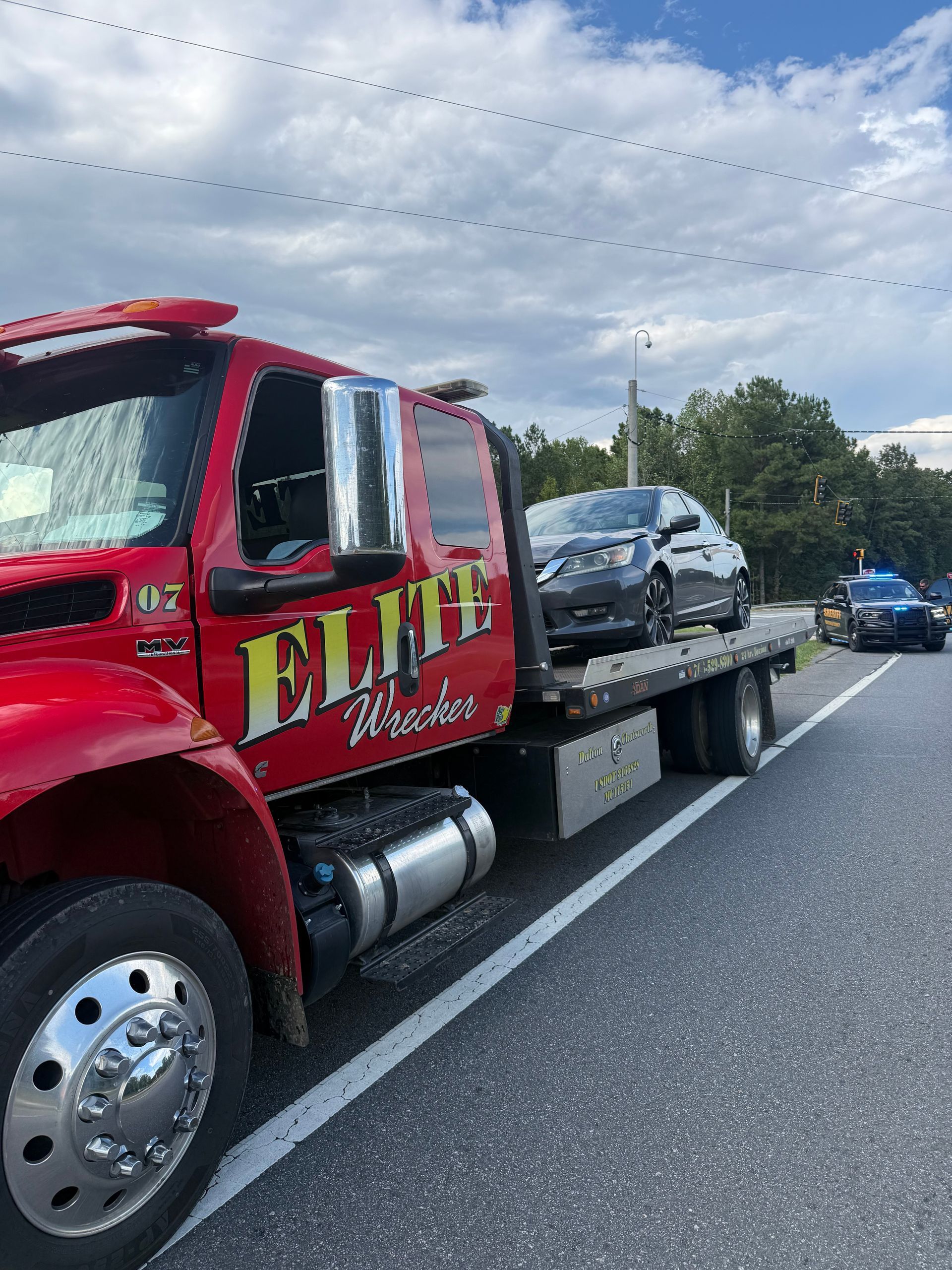 Red tow truck hauling a wrecked silver car on a highway shoulder under a cloudy sky