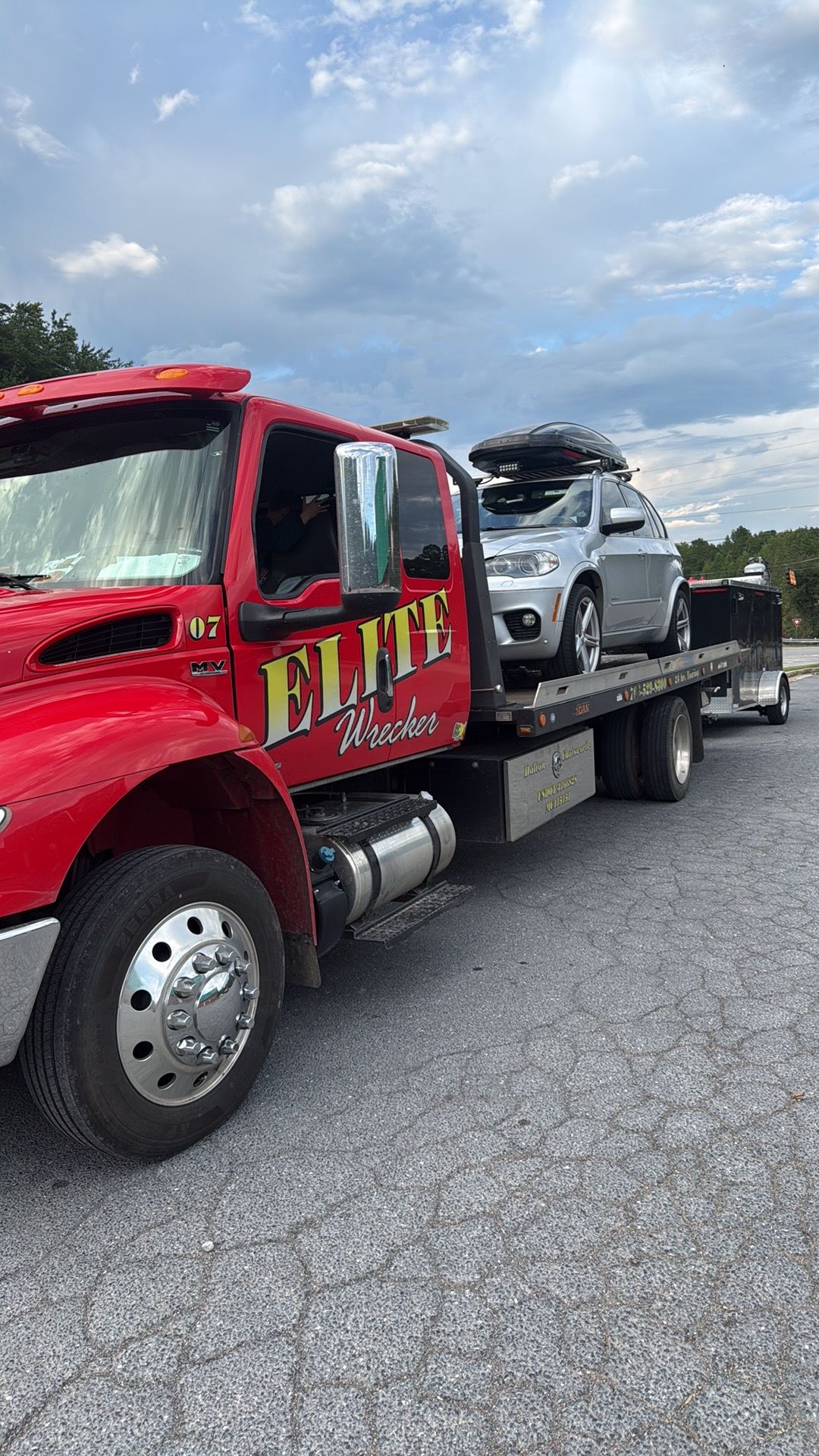 Red tow truck hauling a silver car on a flatbed along a gravel roadside under a cloudy sky