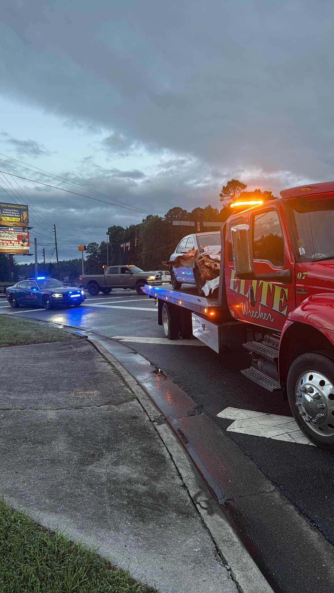 Tow truck with flashing lights towing a blue car on a wet roadside at dusk