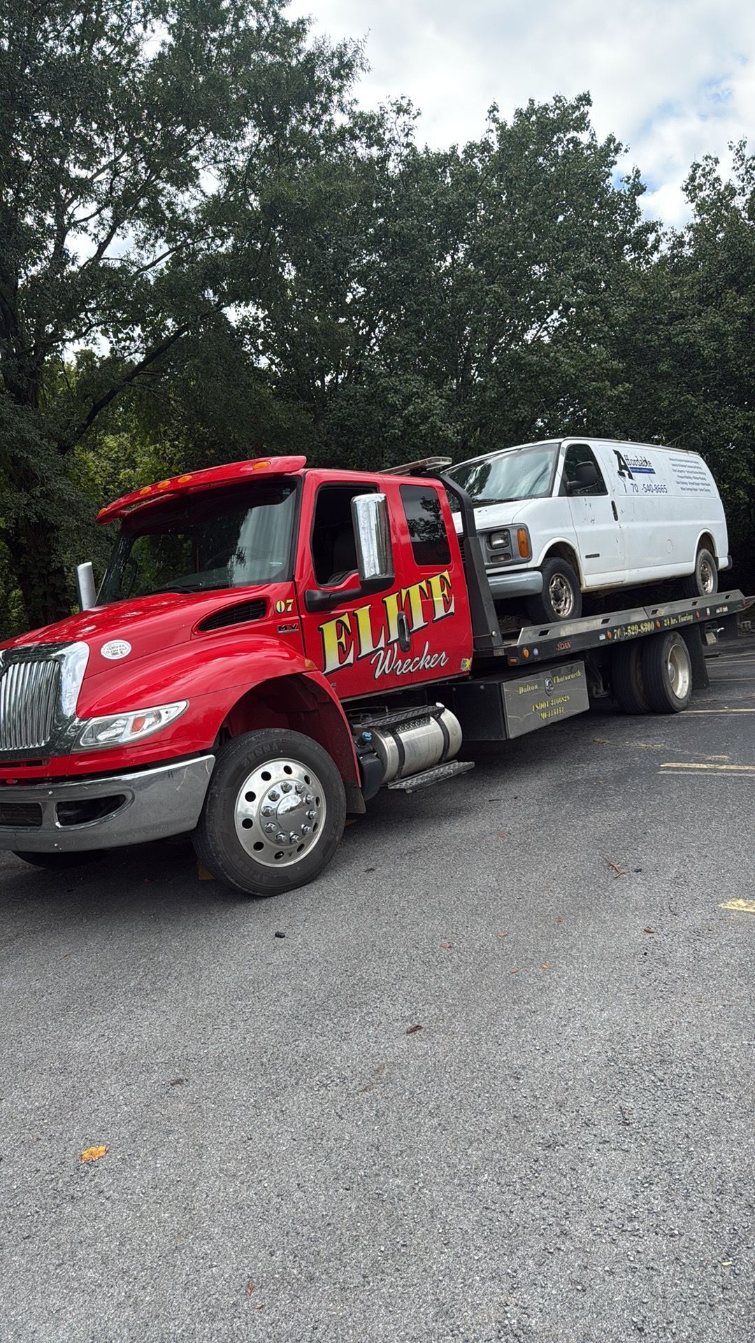 Red tow truck carrying a white cargo van on a flatbed, parked beside a wooded roadside.