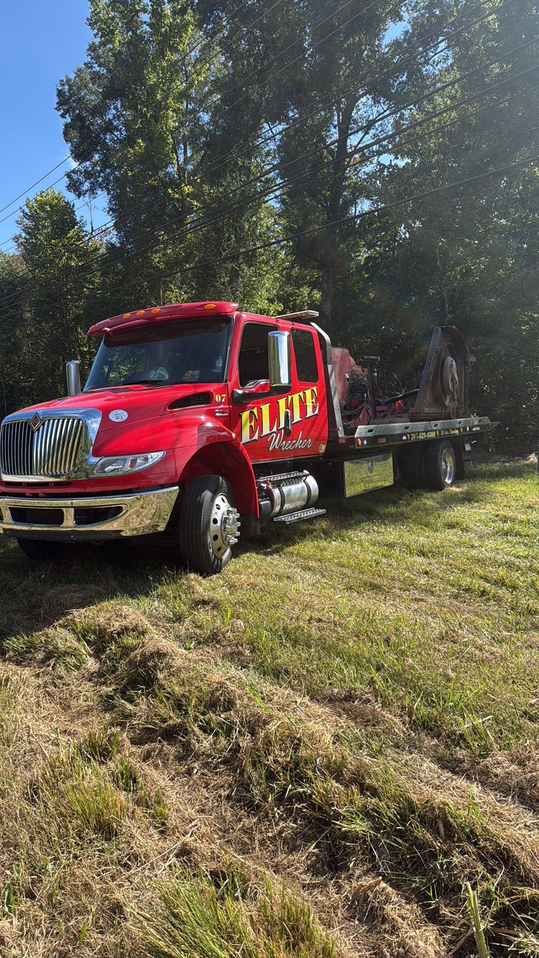 Red flatbed tow truck on a grassy roadside in a sunny wooded area