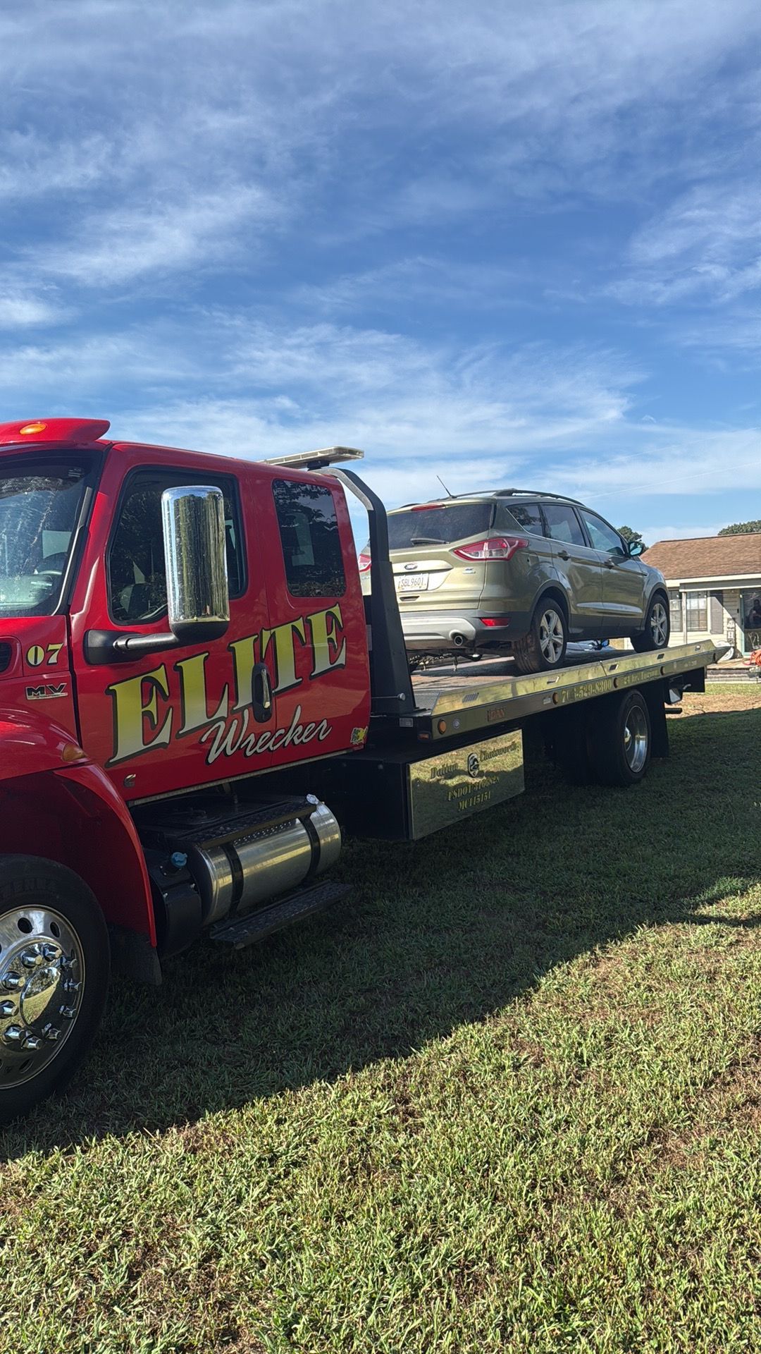 Red flatbed tow truck carrying a gray SUV on a grassy roadside under a blue sky