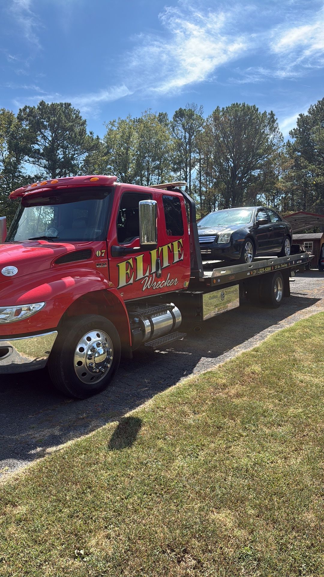 Red tow truck carrying a black car on a flatbed along a roadside under a blue sky