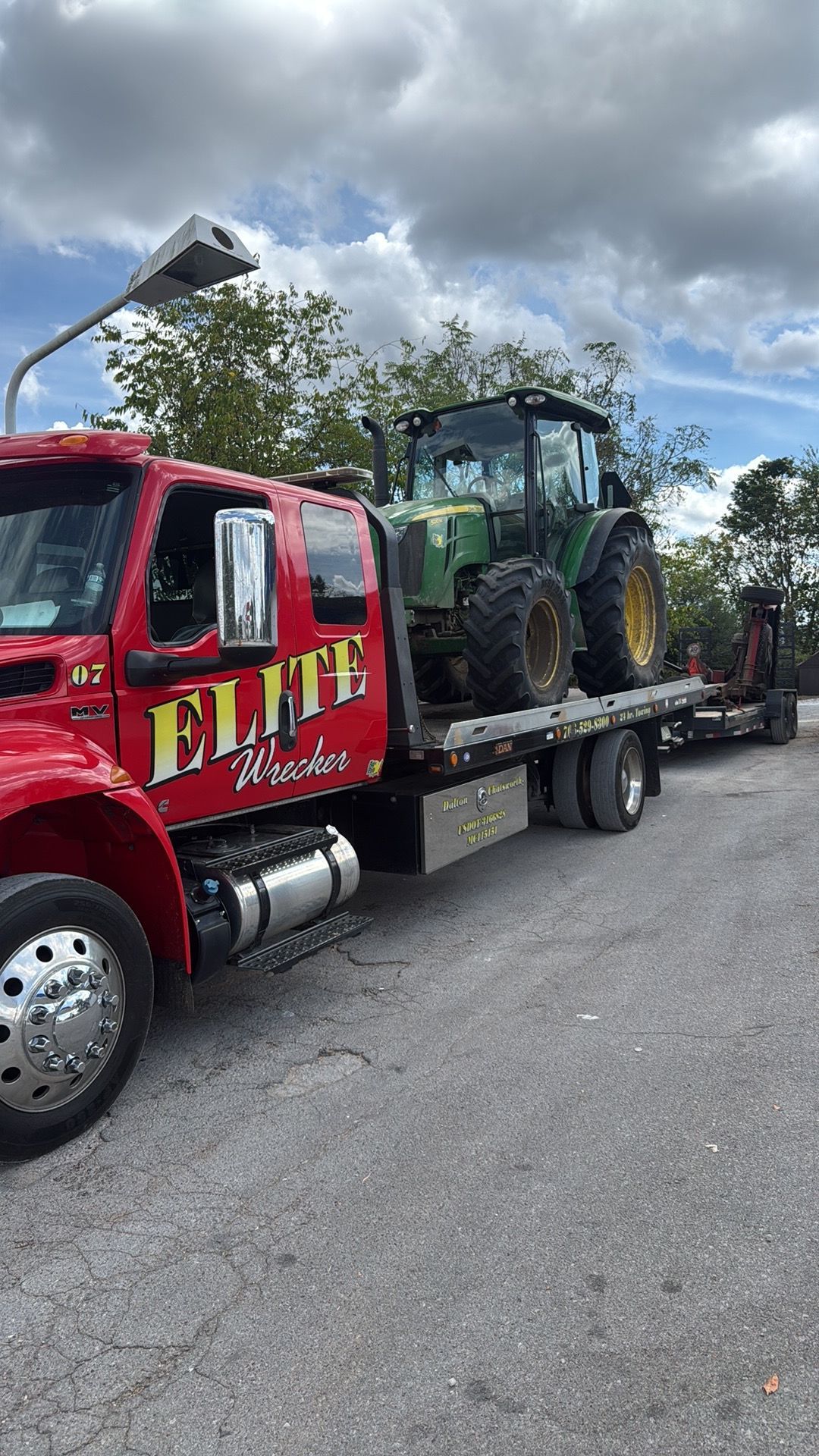 Tow truck carrying a red tractor on a flatbed beside a cloudy roadside.