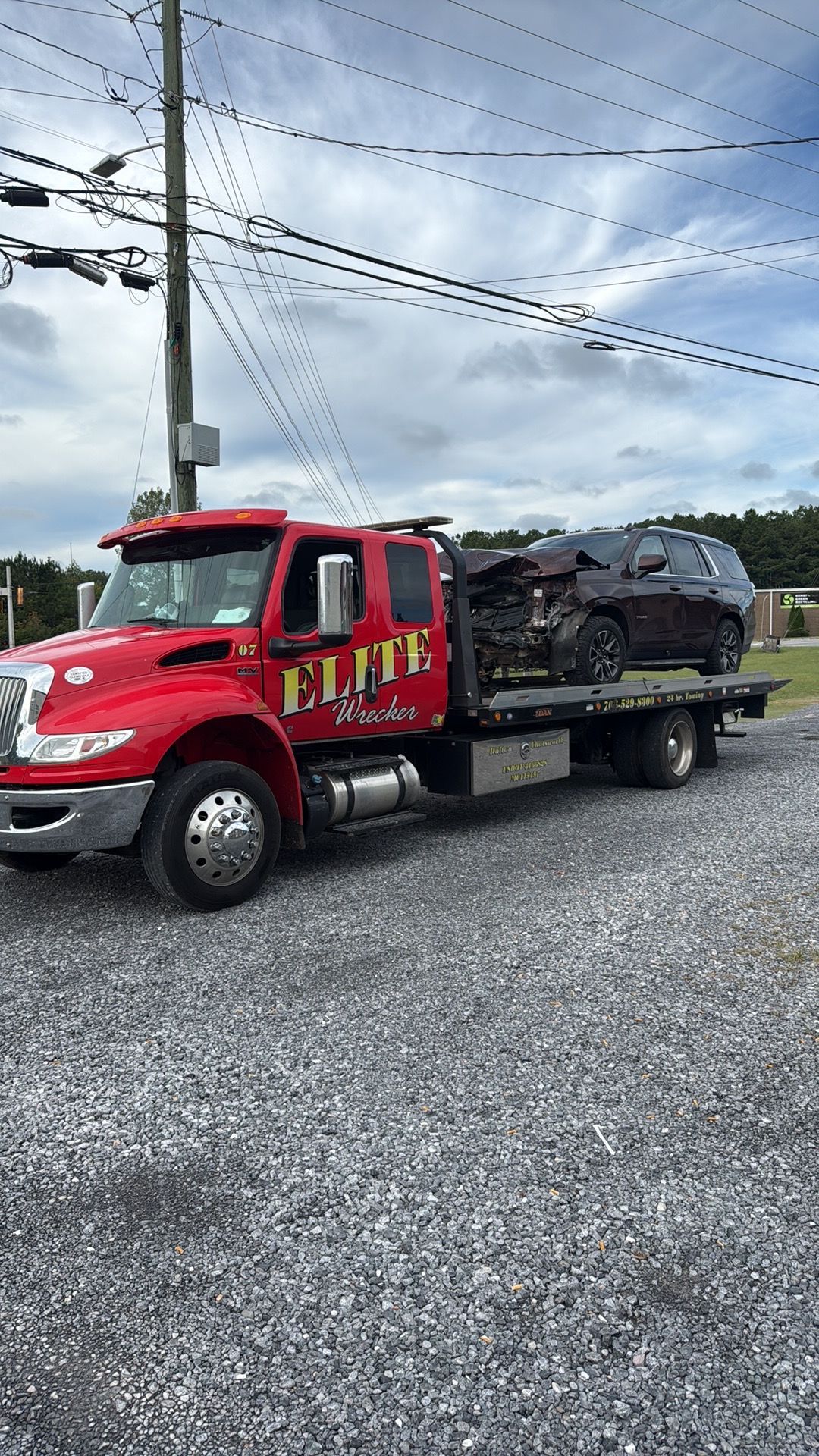 Red tow truck on gravel lot carrying a damaged vehicle under a cloudy sky