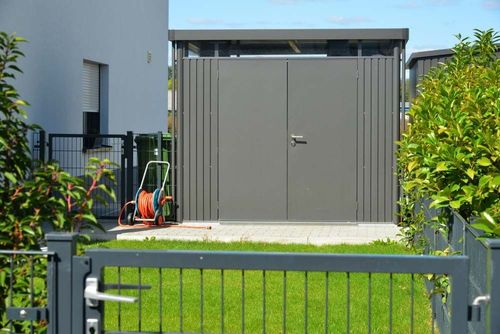 Gray garden shed behind a black metal fence, with a green lawn and orange lawn tools nearby