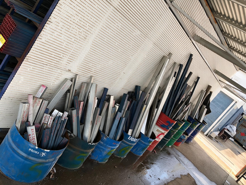 Blue buckets filled with long metal rods and pipes leaning against a chain-link fence in an industrial area