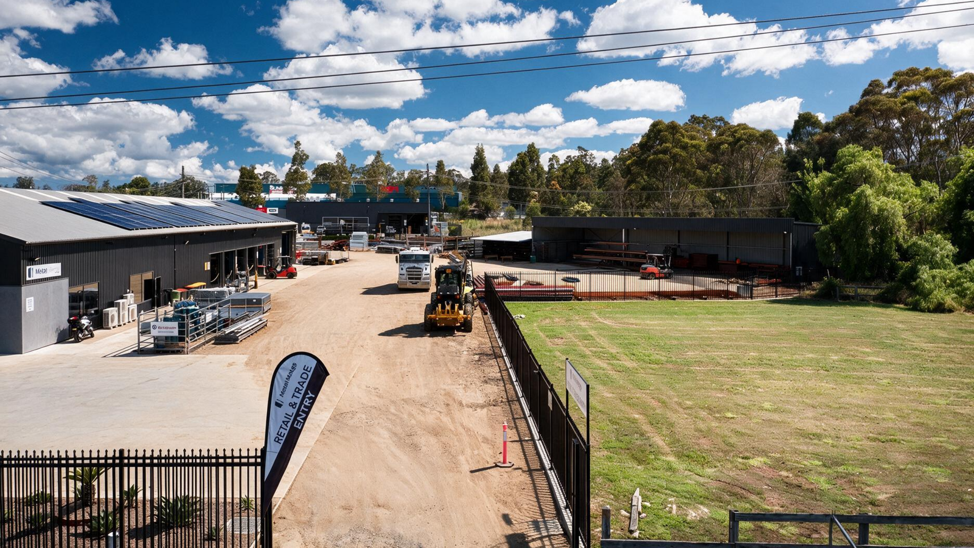 Construction yard with trucks, a small loader, and a grassy field under a cloudy blue sky