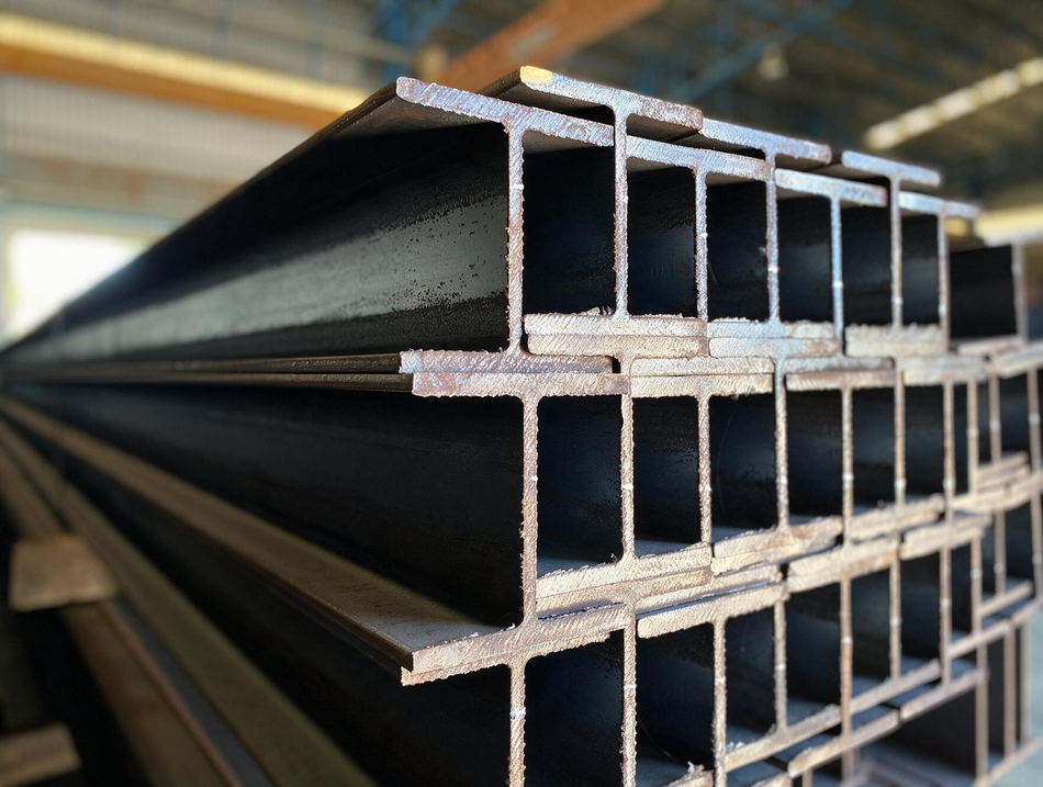 Stacked steel I-beams in a warehouse, showing dark metal cross-sections.