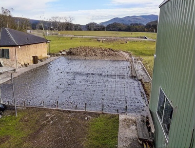 Water-filled rectangular construction pit beside a green building and rural field under cloudy sky