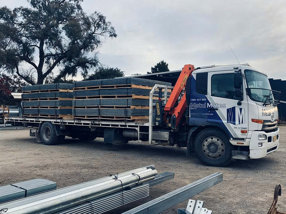 White flatbed truck loaded with metal pipes parked outdoors on a gravel lot
