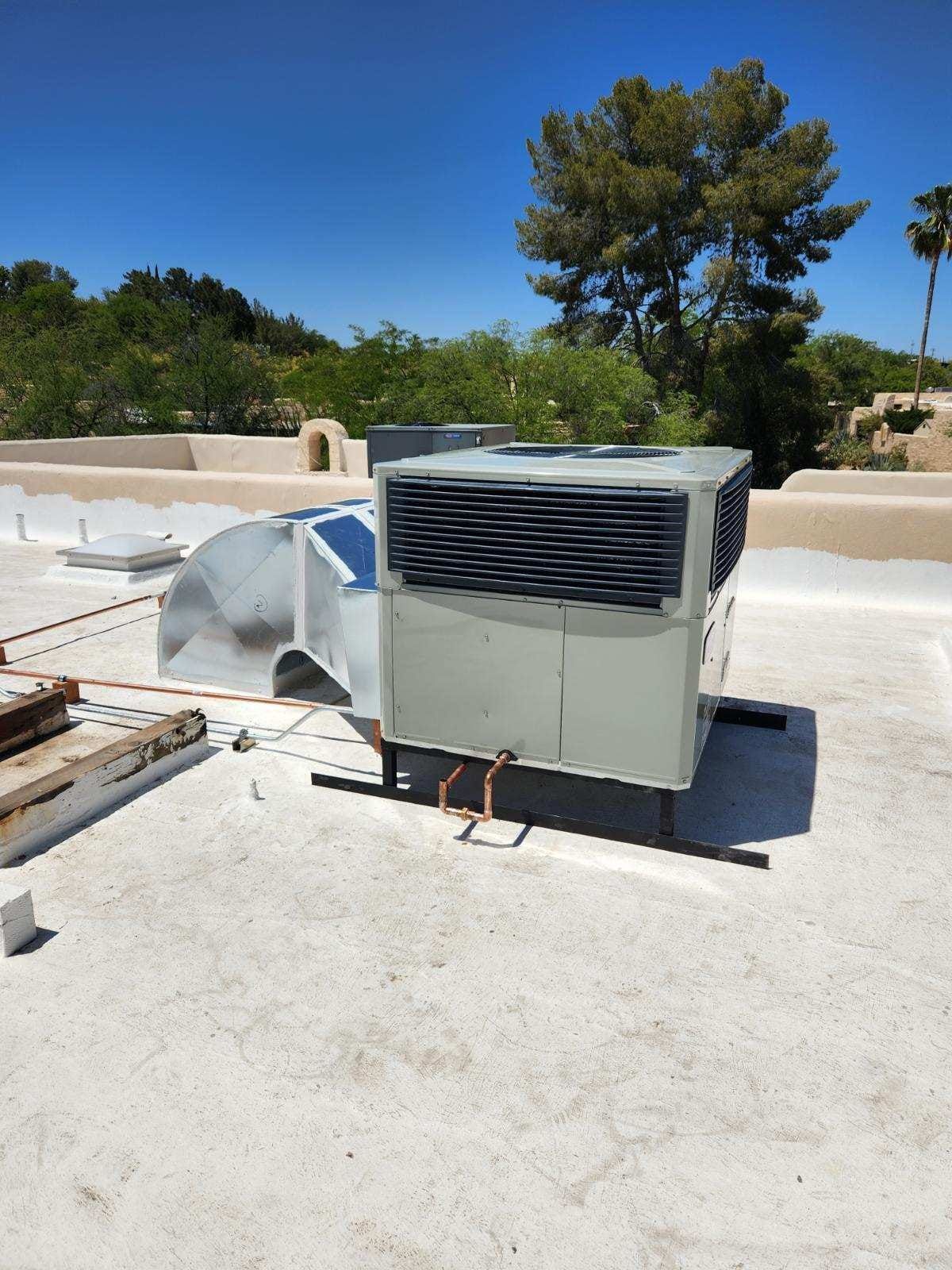 Rooftop HVAC unit with metal ducting under a blue sky, near trees and a low parapet wall