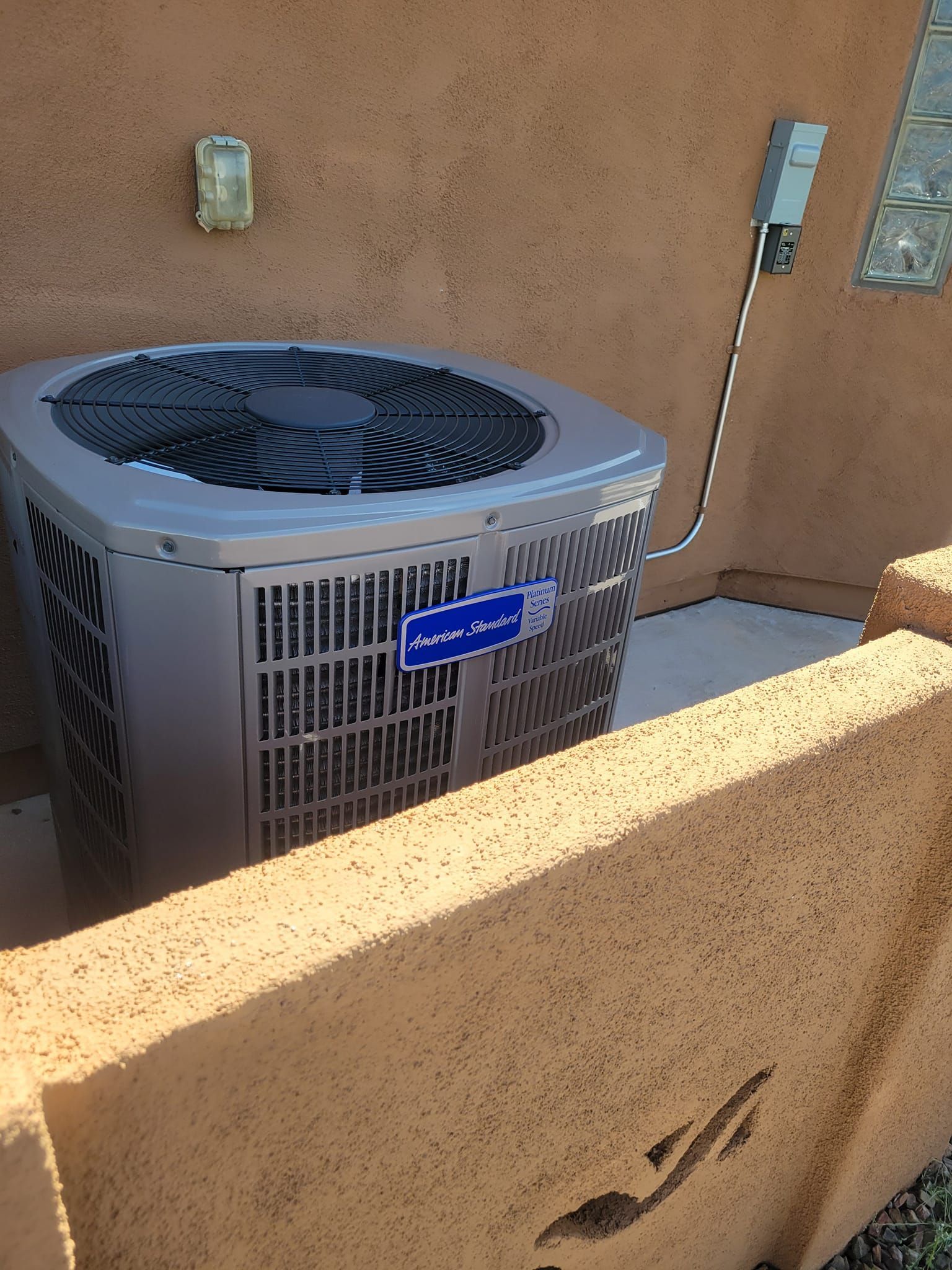 Outdoor HVAC unit beside a stucco wall and fence in bright sunlight