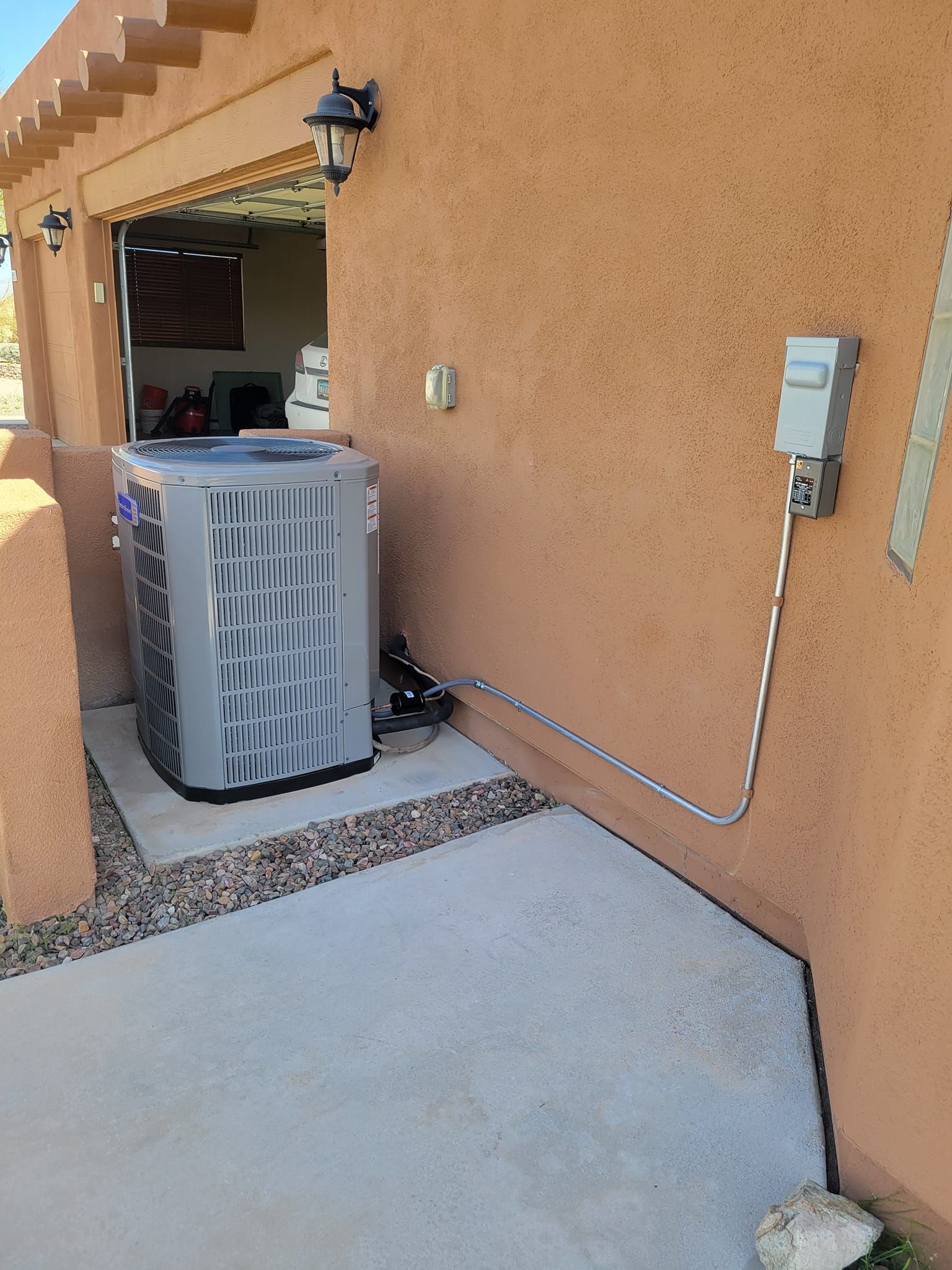 Outdoor HVAC unit beside a tan stucco house, with an open garage and concrete patio.
