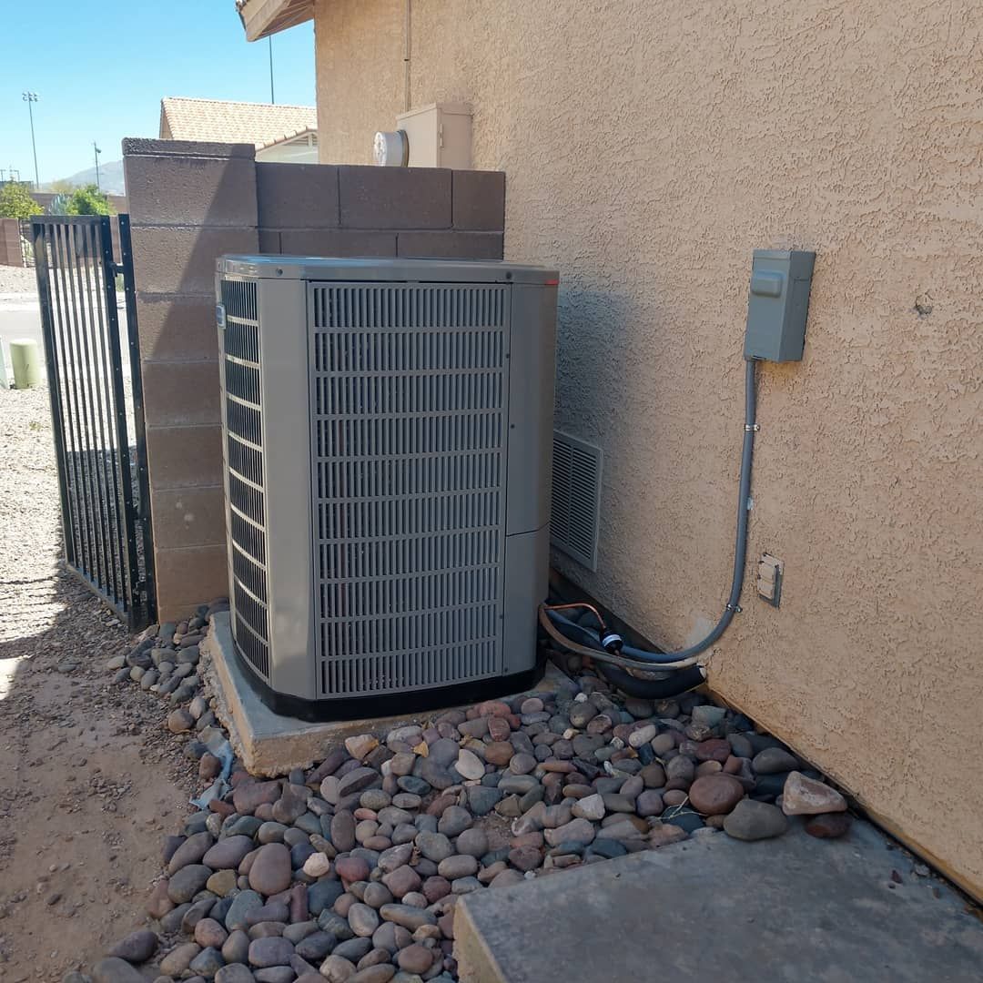 Outdoor air conditioner unit beside a stucco house wall, with electrical box and rocky landscaping.