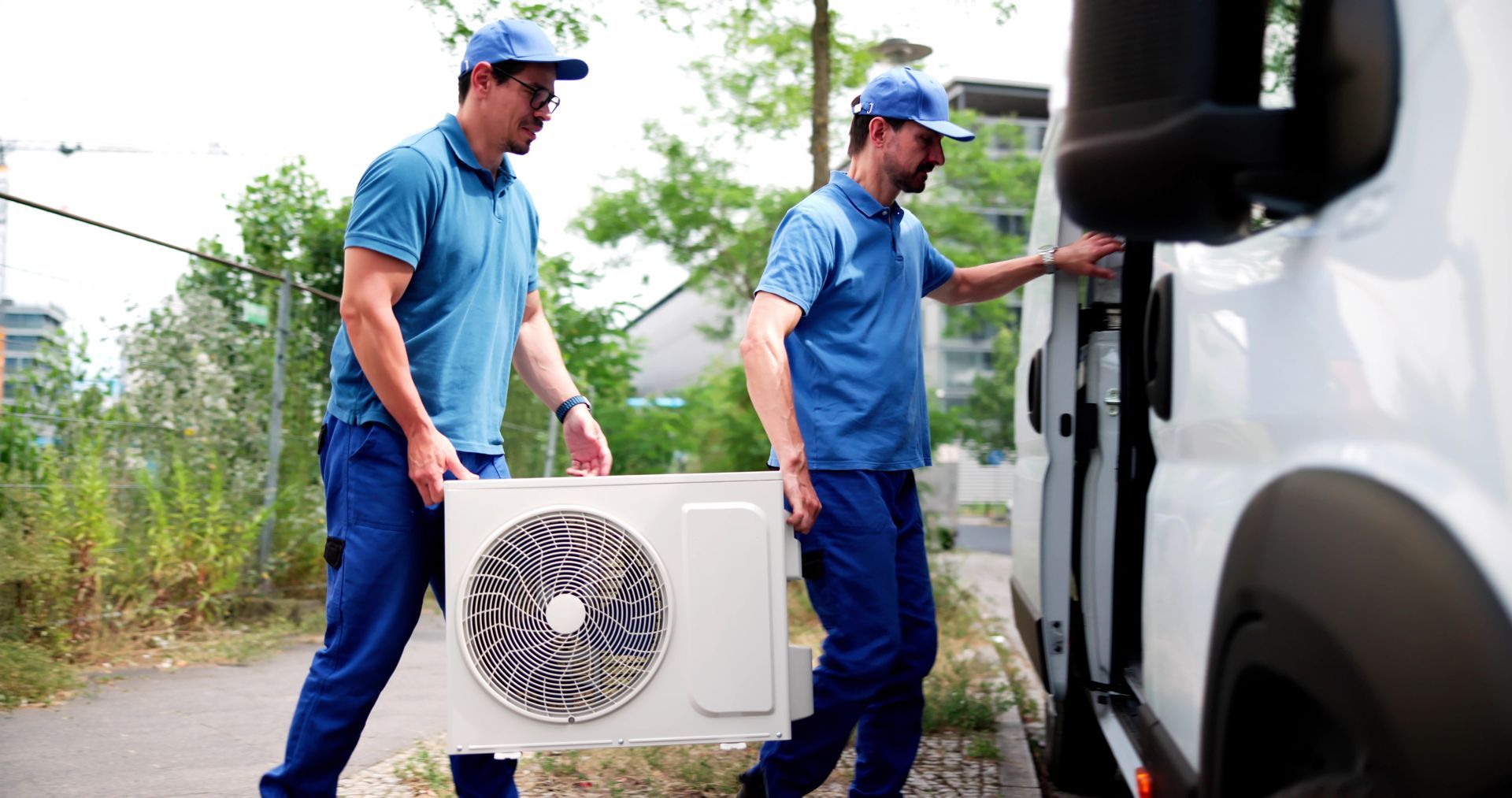 Two workers in blue uniforms carry an air conditioner unit toward a white van outdoors.