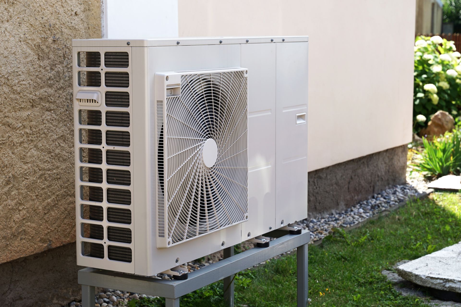 A beige outdoor heat pump unit standing on a metal frame next to a building wall with grass in the background.