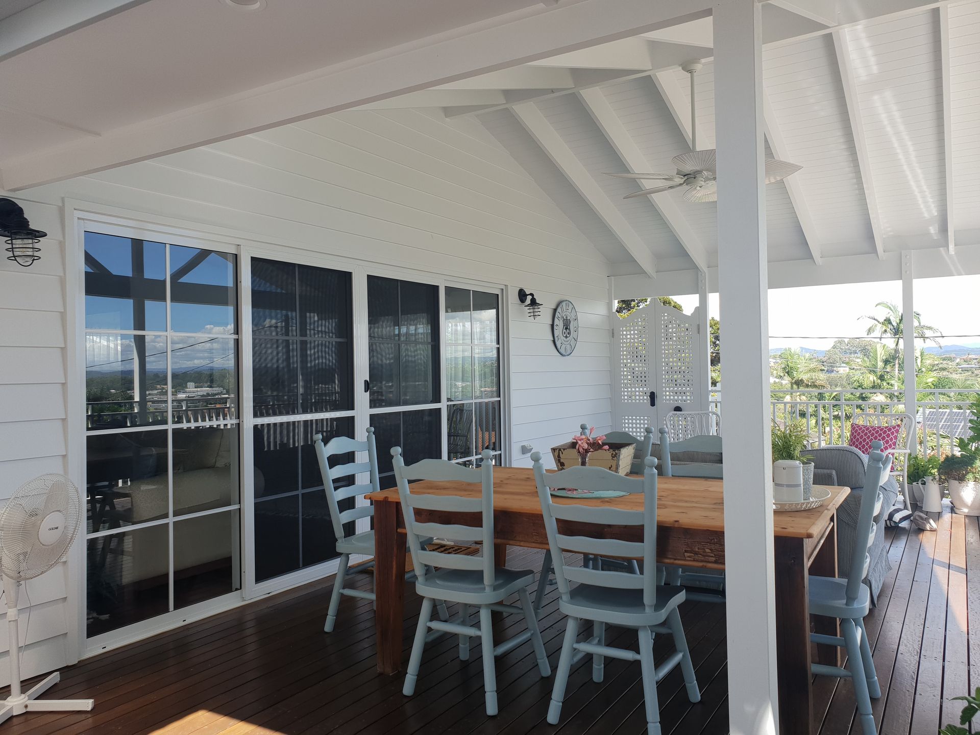 Covered patio with wooden dining table, white chairs, and sliding glass doors overlooking a garden.