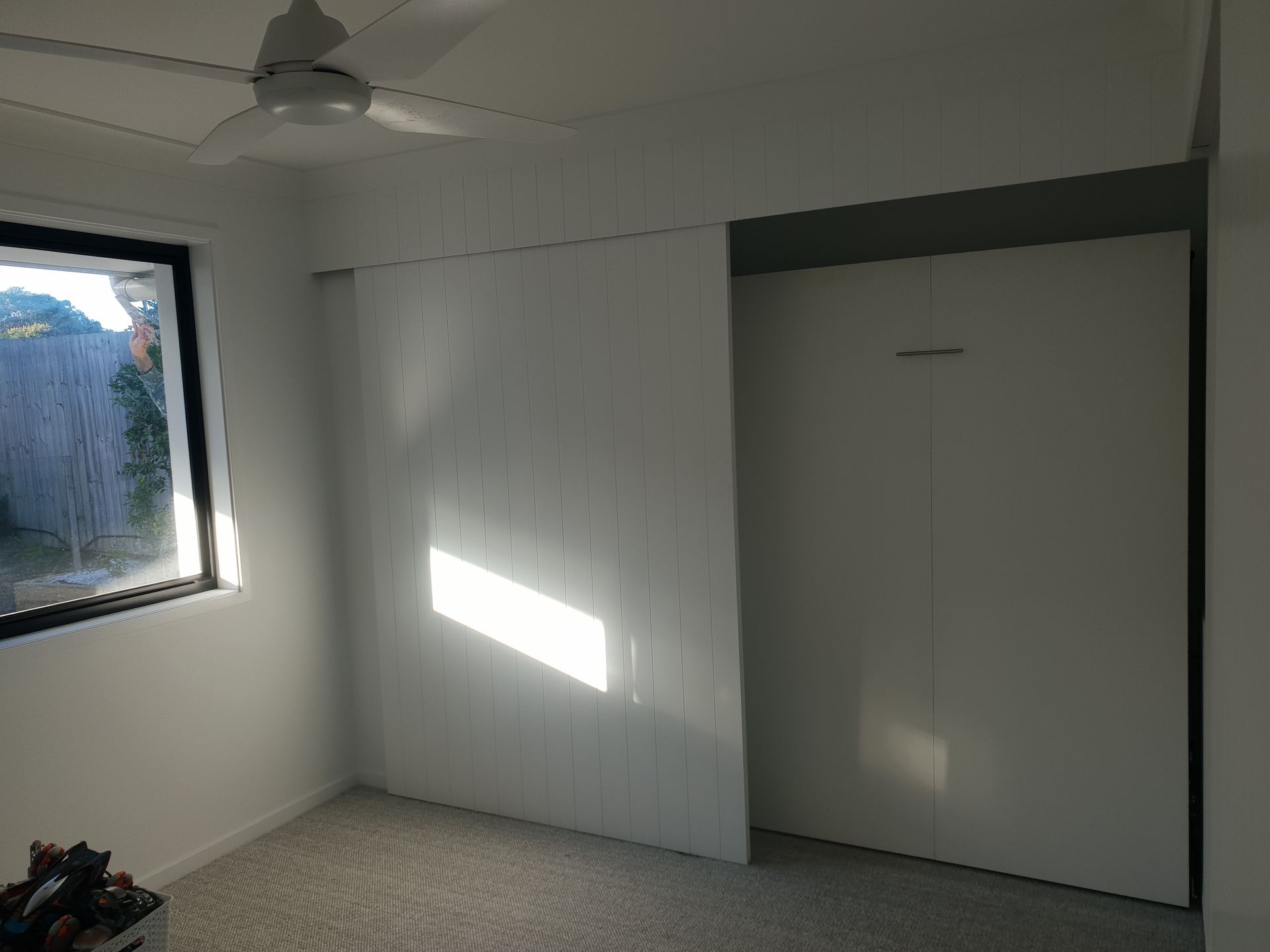 Empty white room with window, ceiling fan, and closet door; sunlight on the wall.