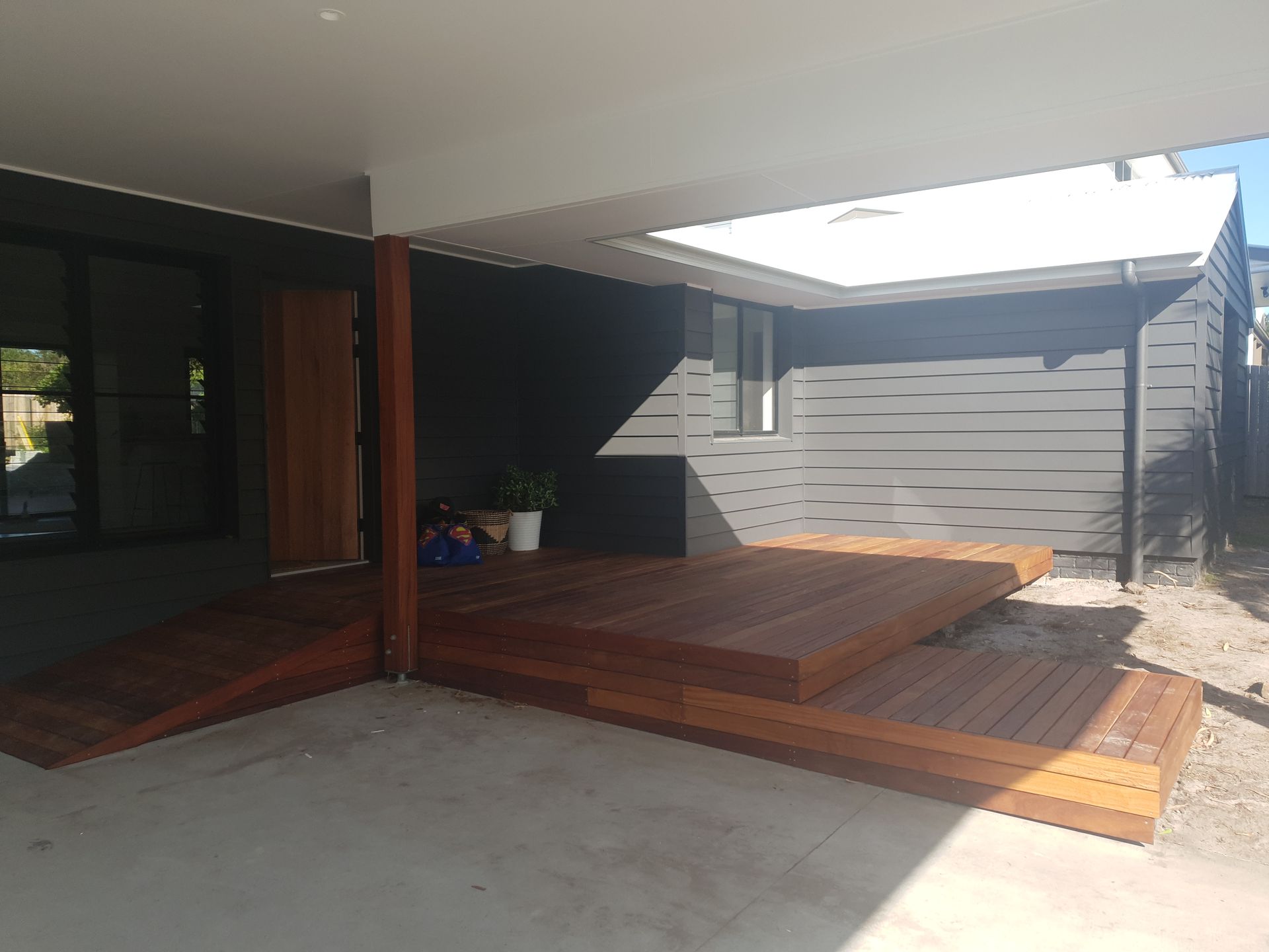 Covered wooden deck beside a modern house, with gray siding and a concrete patio in the foreground