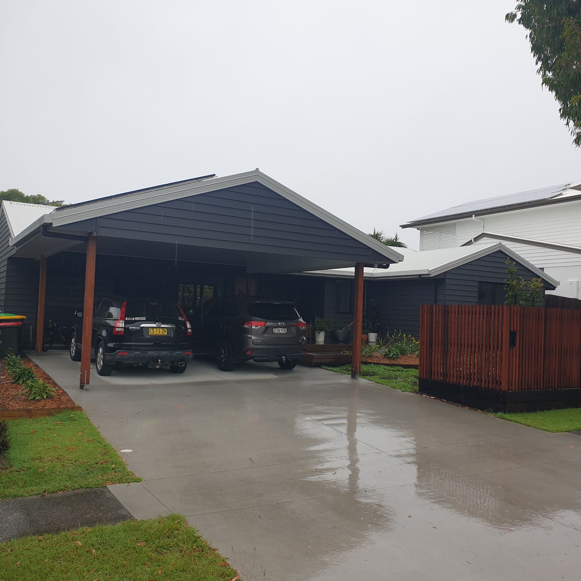 Rainy driveway with two cars parked under a covered carport beside a white house and wooden fence.