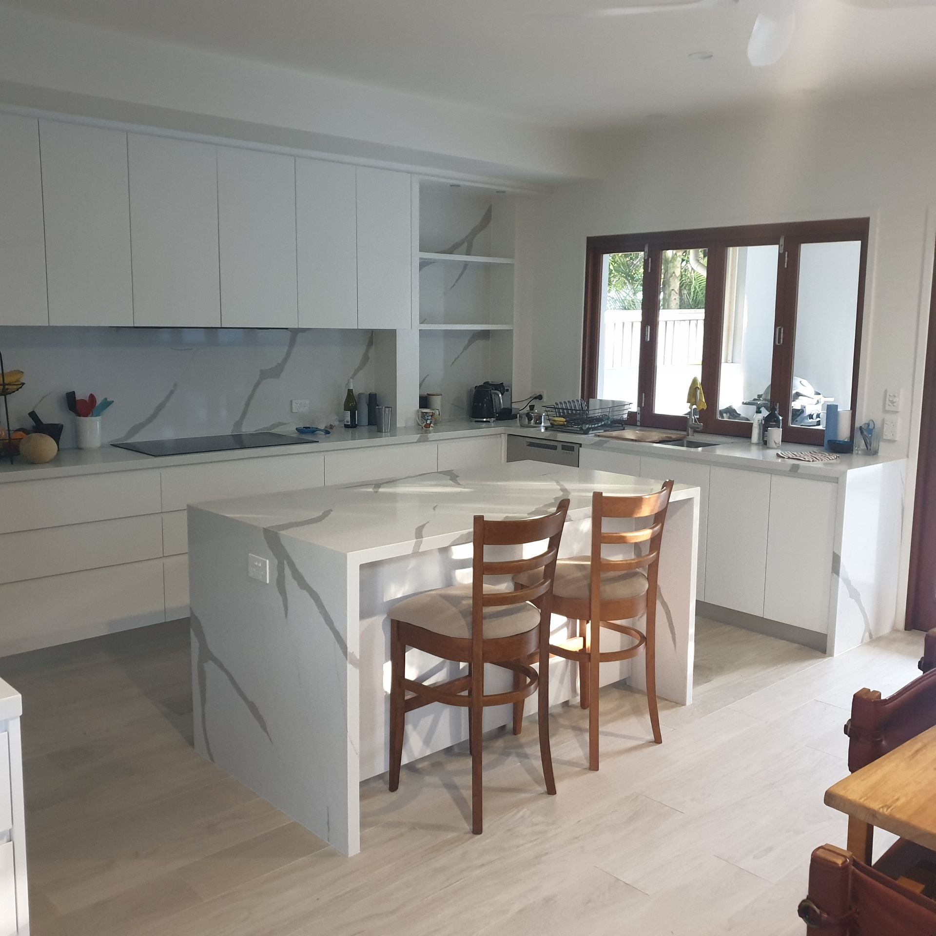 Bright white modern kitchen with an island, wooden chairs, and large windows letting in daylight