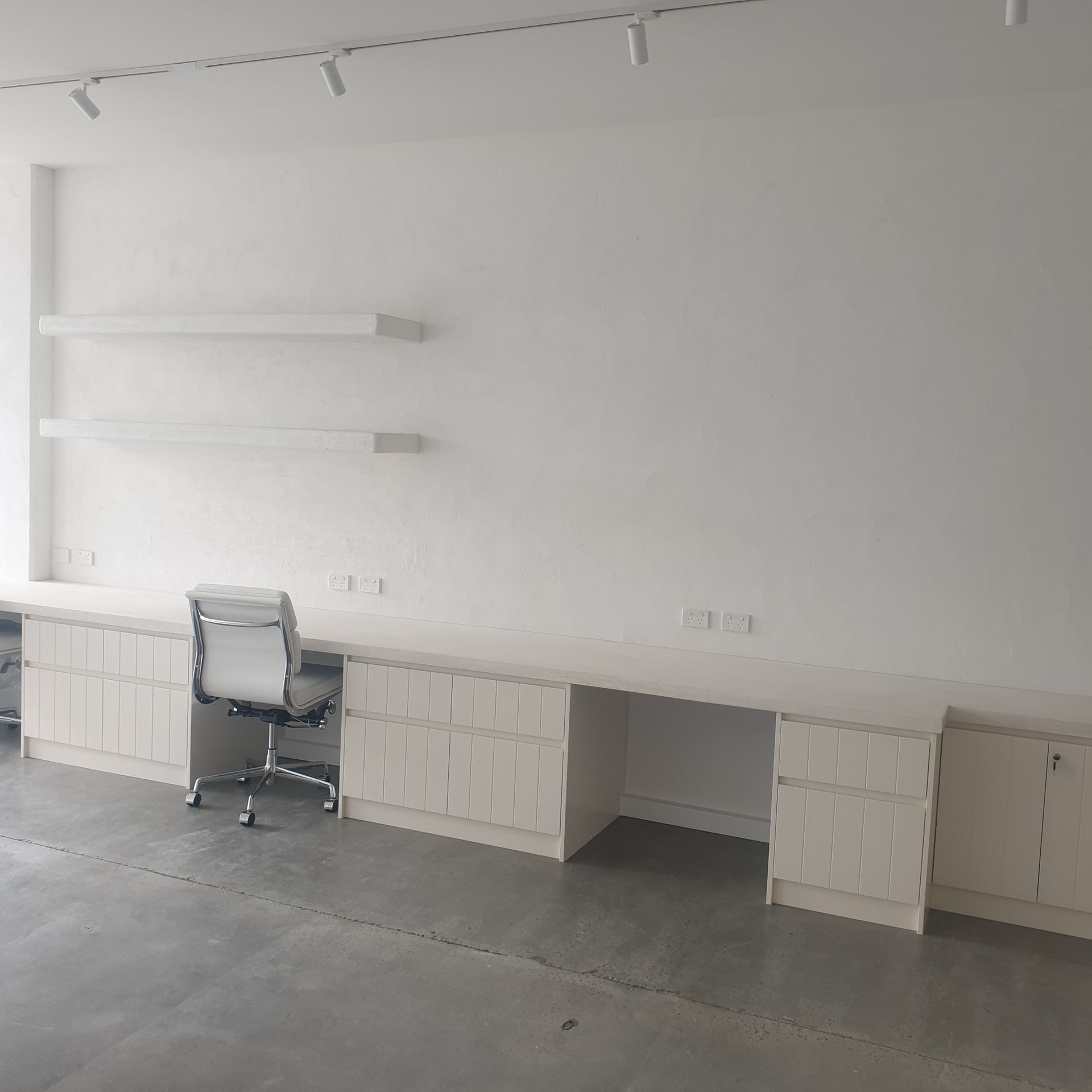 Empty white office with a desk, swivel chair, wall shelves, and a concrete floor.