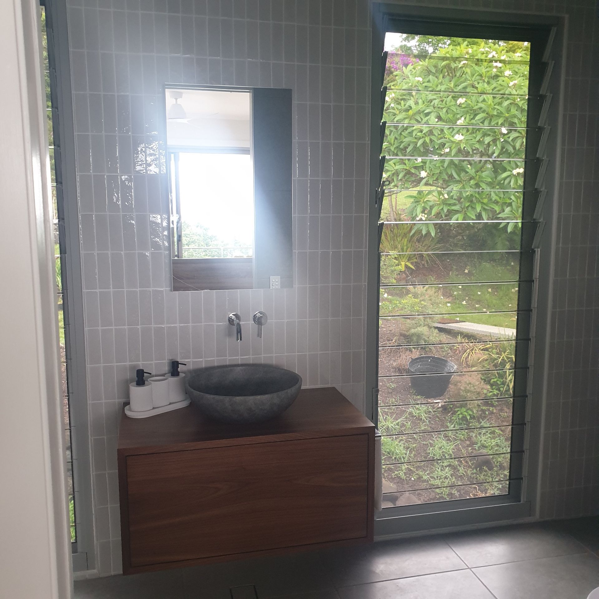 Modern bathroom vanity with wood cabinet, stone sink, mirror, and glass door opening to greenery