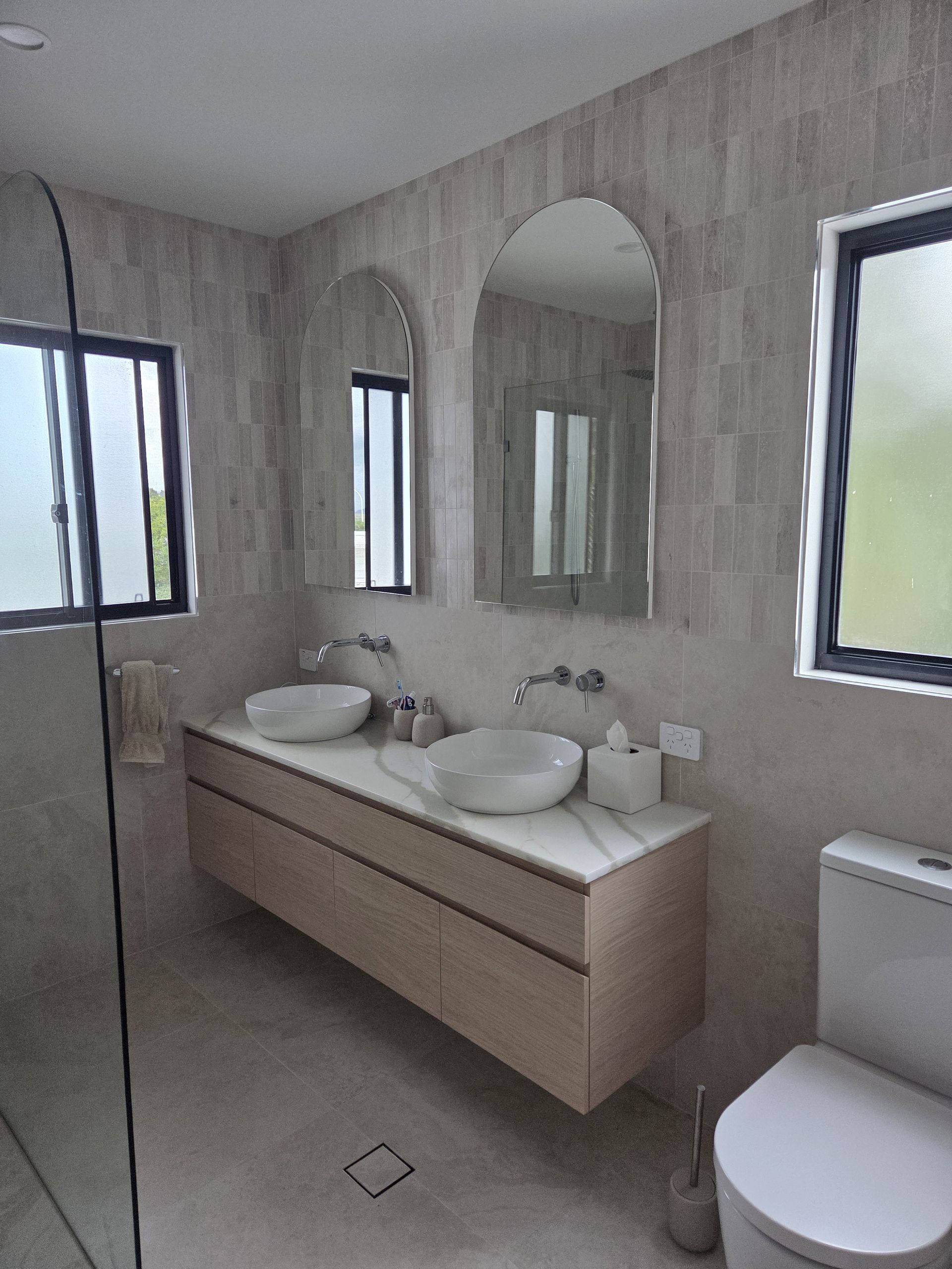 Modern bathroom with double-sink vanity, large mirror, windows, and a toilet.