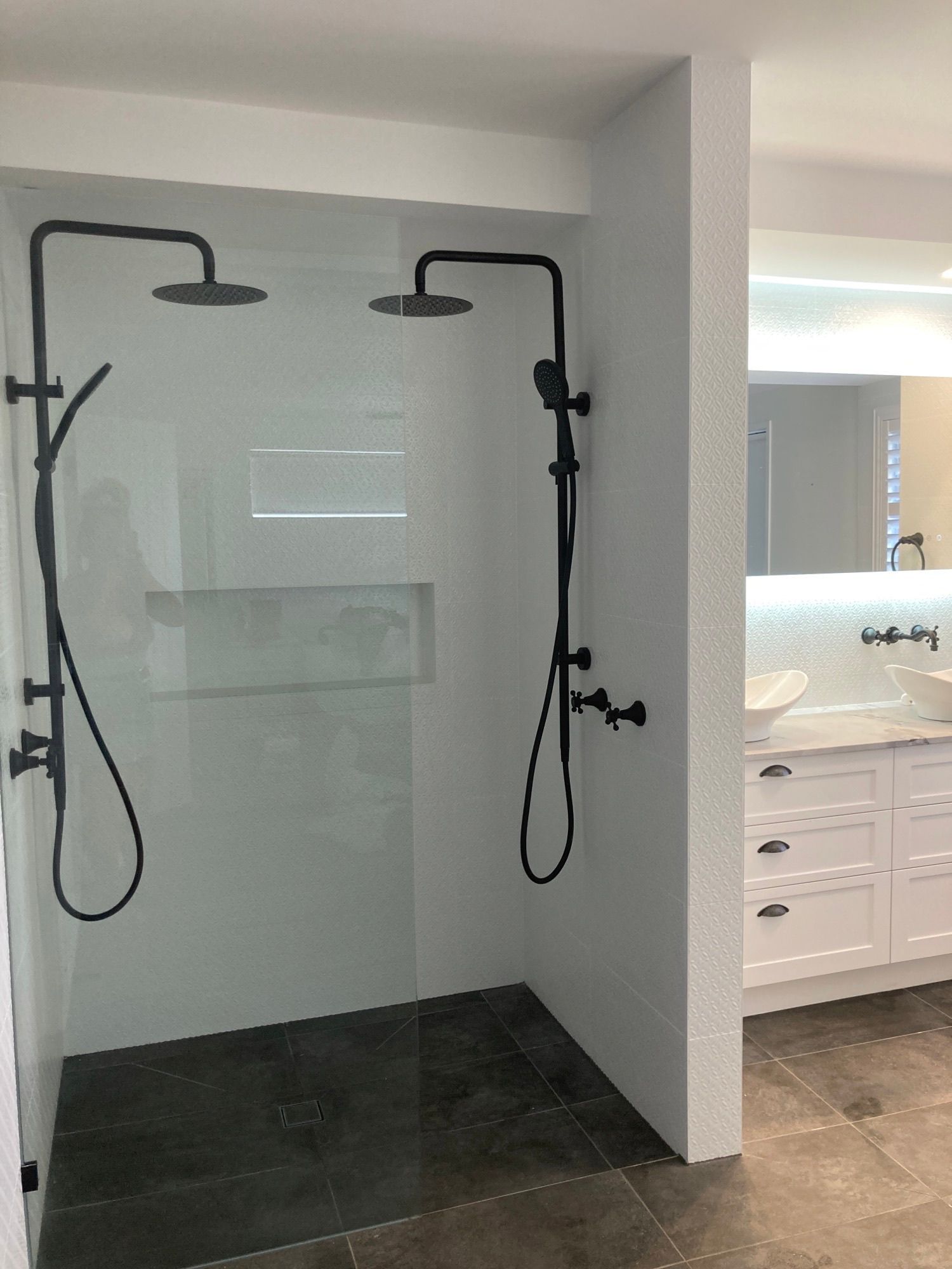Bathroom shower with two black handheld showerheads and a tiled floor next to a vanity sink.