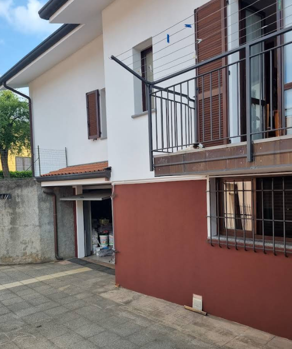 Vista laterale di una casa a due piani. Esterno bianco e rosso mattone, persiane marroni, piccolo balcone, porta del garage aperta.