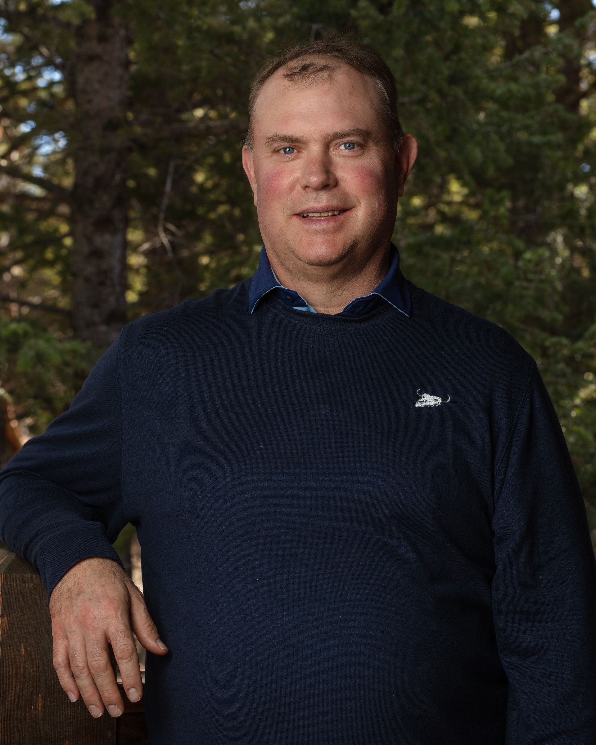 A man in a blue sweater is standing in front of trees.