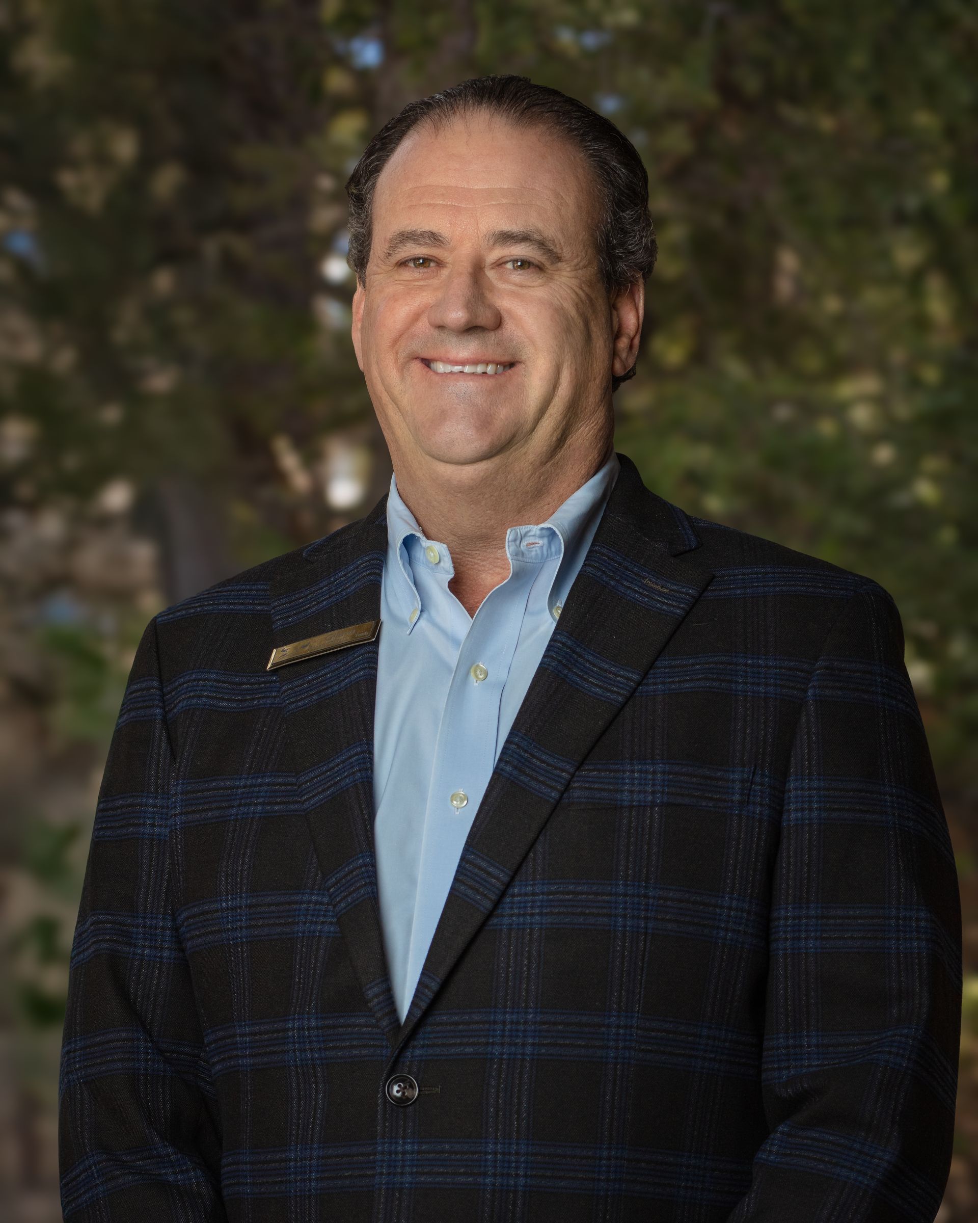 A man in a suit and blue shirt is standing in front of trees.