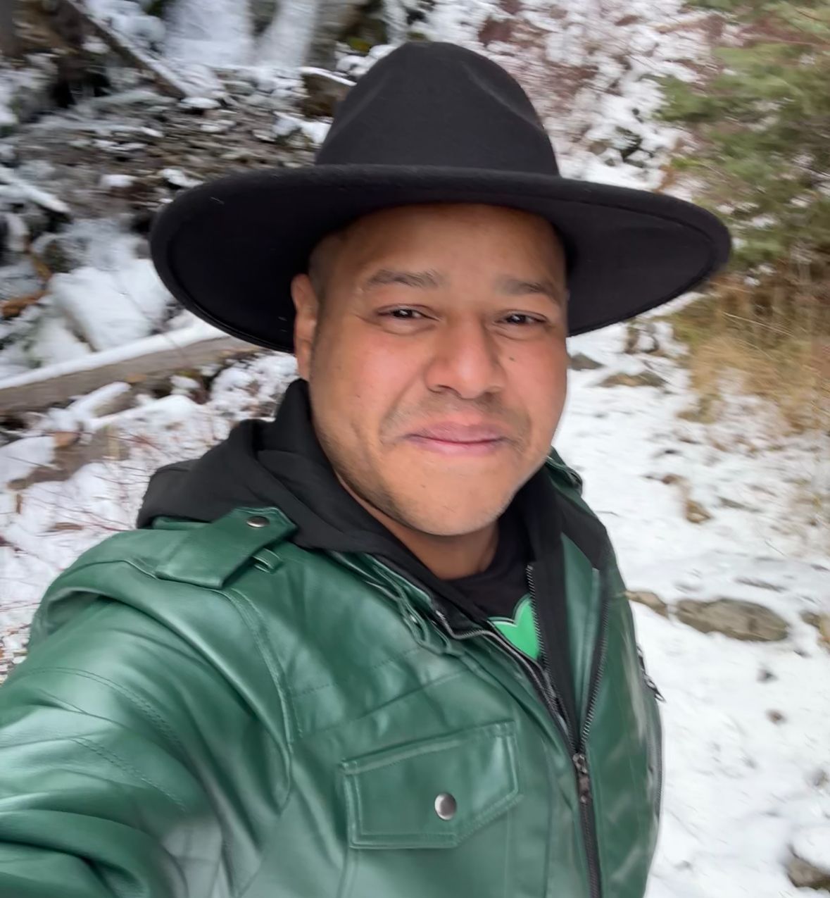 A man wearing a black hat and a green jacket is taking a selfie in the snow.