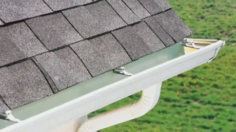 Close-up of a roof with gray shingles and a white gutter. A downspout curves down towards green grass.