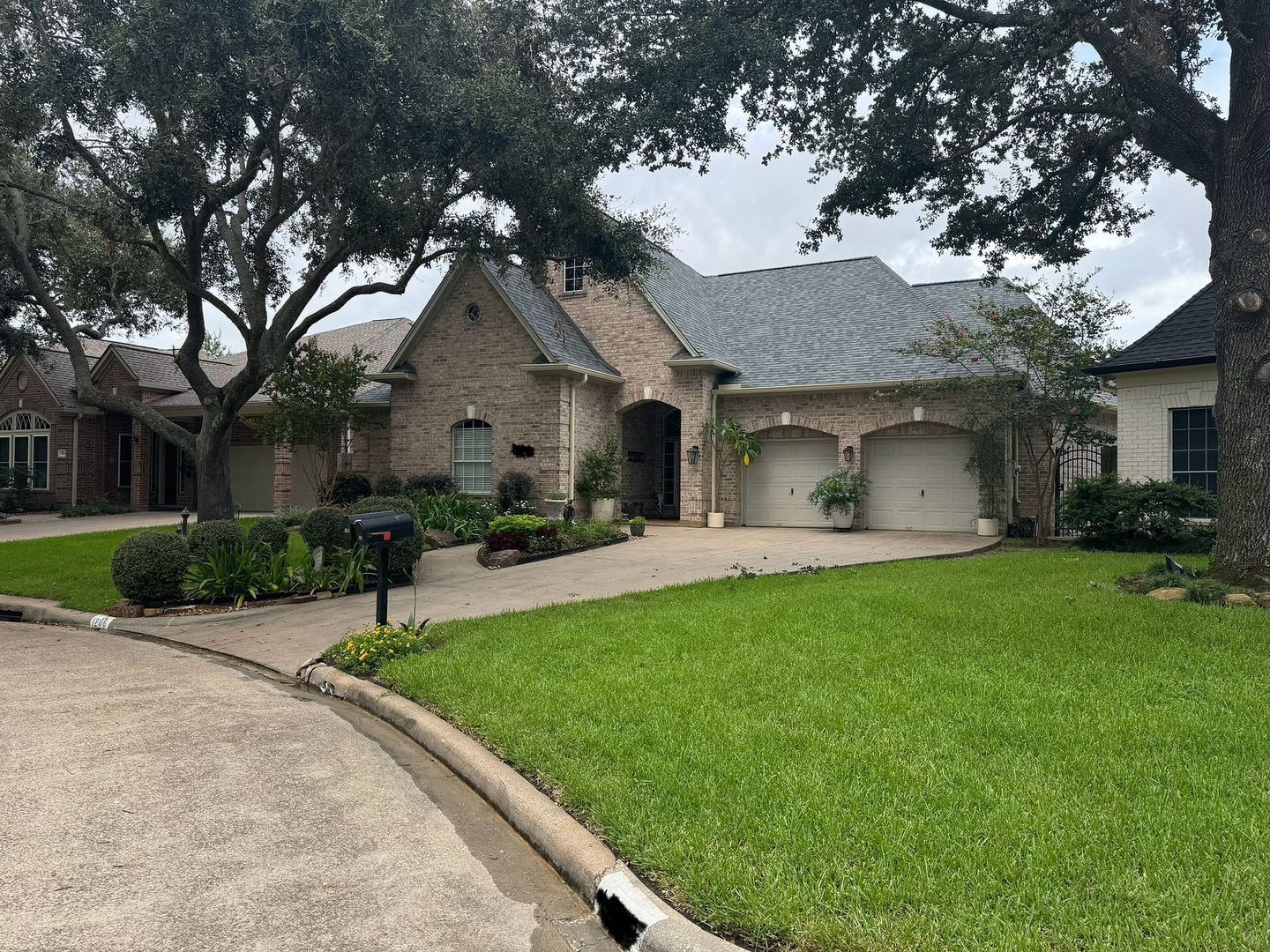 House with a brick exterior, a gray roof, and a two-car garage. Green lawn with trees.