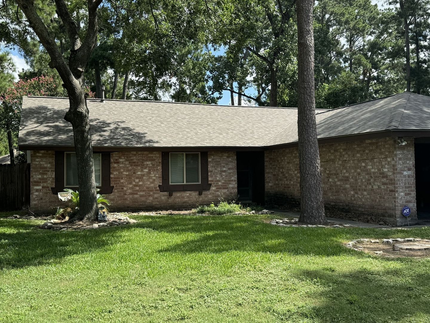 Brick house with brown roof, surrounded by trees, and green grass in front.