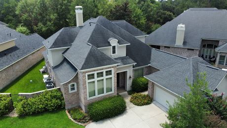 House with dark roof and chimney, brick and light siding, surrounded by green trees and grass.