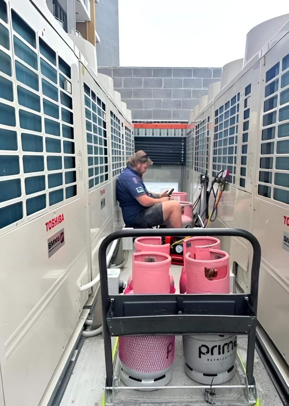 Man seated, working between two large AC units. Cart with pink and grey propane tanks  — Sparky Mid North Coast In Forster, NSW