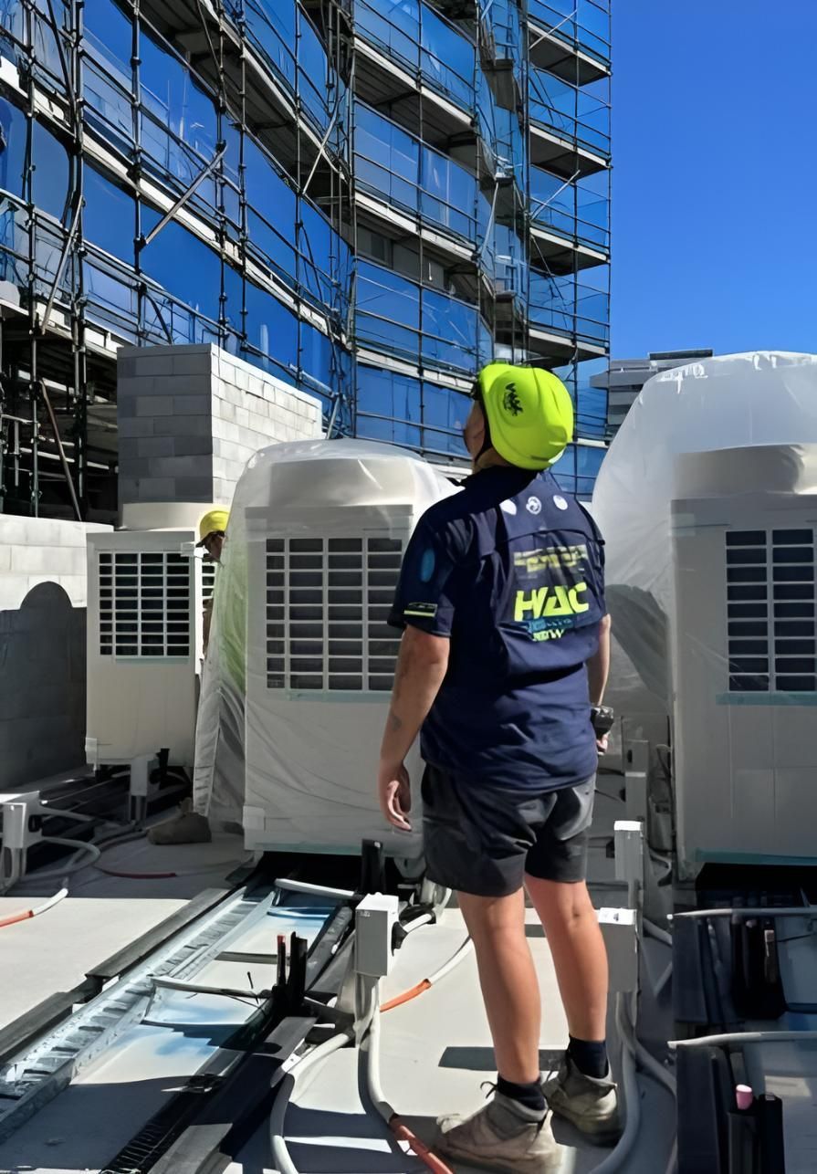A worker in a yellow helmet and uniform inspects HVAC units on a rooftop, construction in the background  — Sparky Mid North Coast In Forster, NSW
