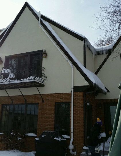 A two-story house with cream-colored stucco, a brick ground floor, and snow-covered roof peaks with icicles.