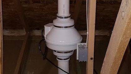 A radon mitigation fan installed in an attic, connected to white PVC pipes and a nearby electrical outlet.
