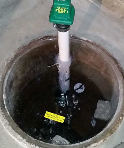 A sump pump submerged in water inside a concrete pit, featuring a white PVC pipe extending upward to a green motor unit.