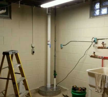 A basement corner with a white sump pump pipe, a metal conduit, a yellow stepladder, and a utility sink.