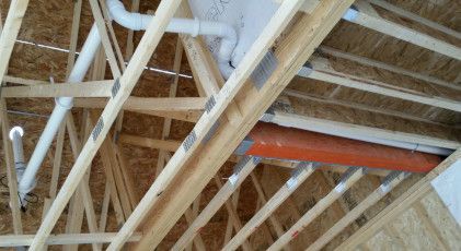Unfinished attic framing showing wood trusses, exposed sheathing, white PVC piping, and an orange HVAC duct.