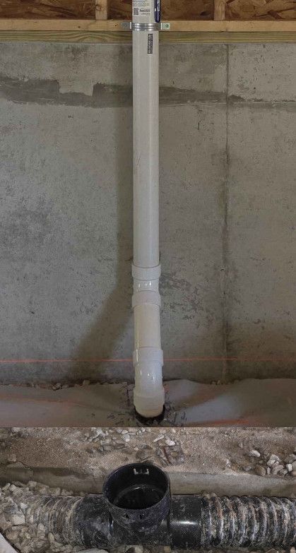 A vertical white PVC pipe attached to a concrete wall, positioned above a black horizontal drainage pipe in a basement.