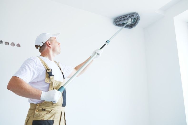 Painter in white uniform rolling paint on a white ceiling with a long-handled roller.