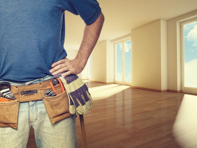 Construction worker in blue shirt with tool belt and gloves, standing in a bright empty room with hardwood floors