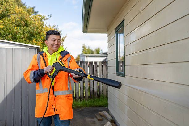 Worker in orange safety gear uses a spray wand beside a house exterior